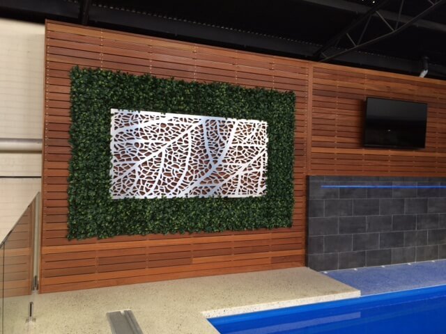 Artificial green wall panel by Designer Plants USA installed at a pool shop, featuring lush faux hedge panels and metal wall art.