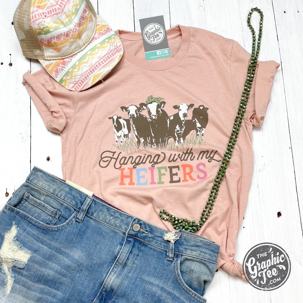 White short-sleeve graphic T-shirt featuring "Hanging With My Heifers" text from The Graphic Tee, displayed on a hanger.