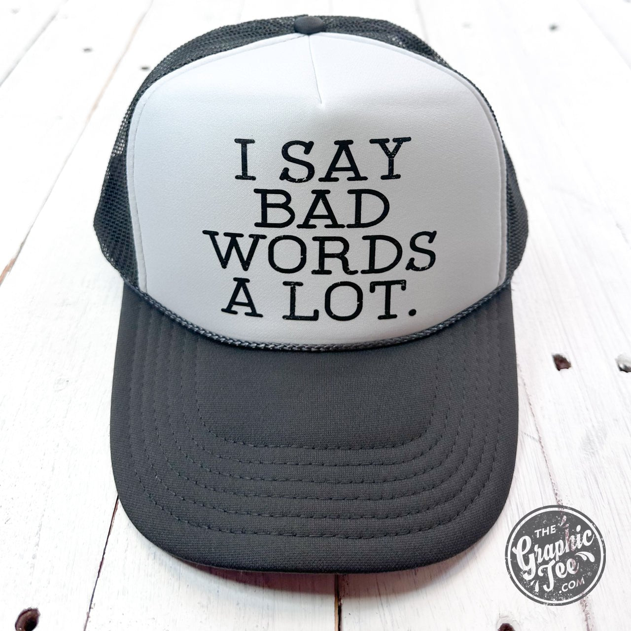 Black foam trucker cap with bold white text saying "I Say Bad Words A Lot" by The Graphic Tee, adjustable snapback style.