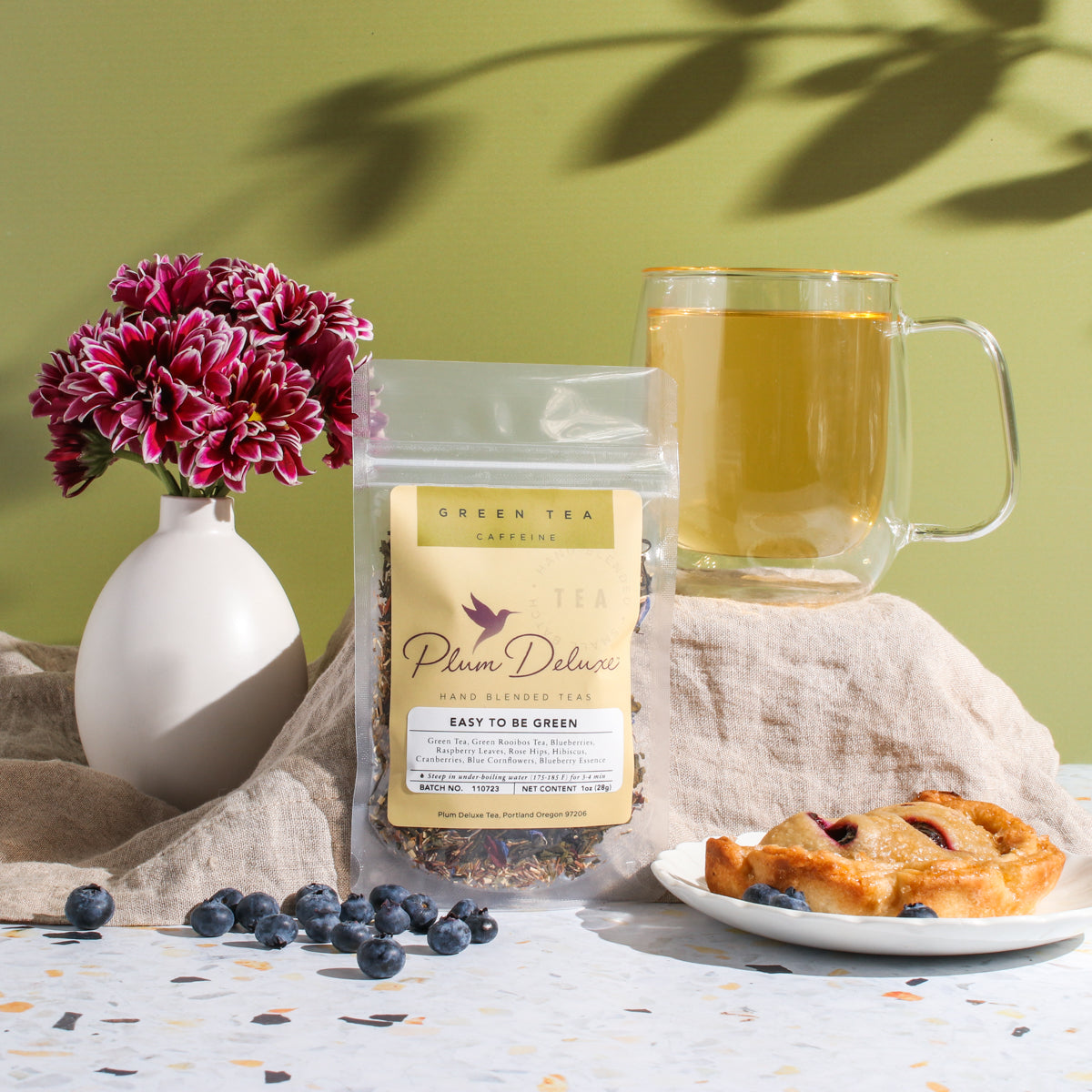 Plum Deluxe Easy to Be Green Tea with Blueberry and Hibiscus in a transparent tea jar showing dried herbs and fruit pieces.