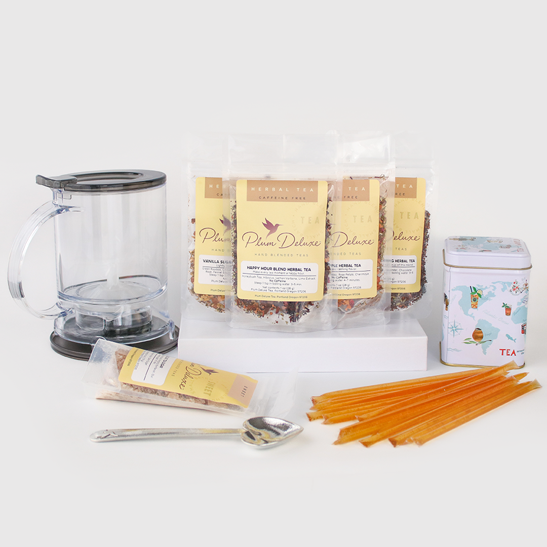 Plum Deluxe Tea Ultimate Hot Tea Bundle with assorted teas, sweets, scoop, tin, and tea maker arranged on a wooden surface.