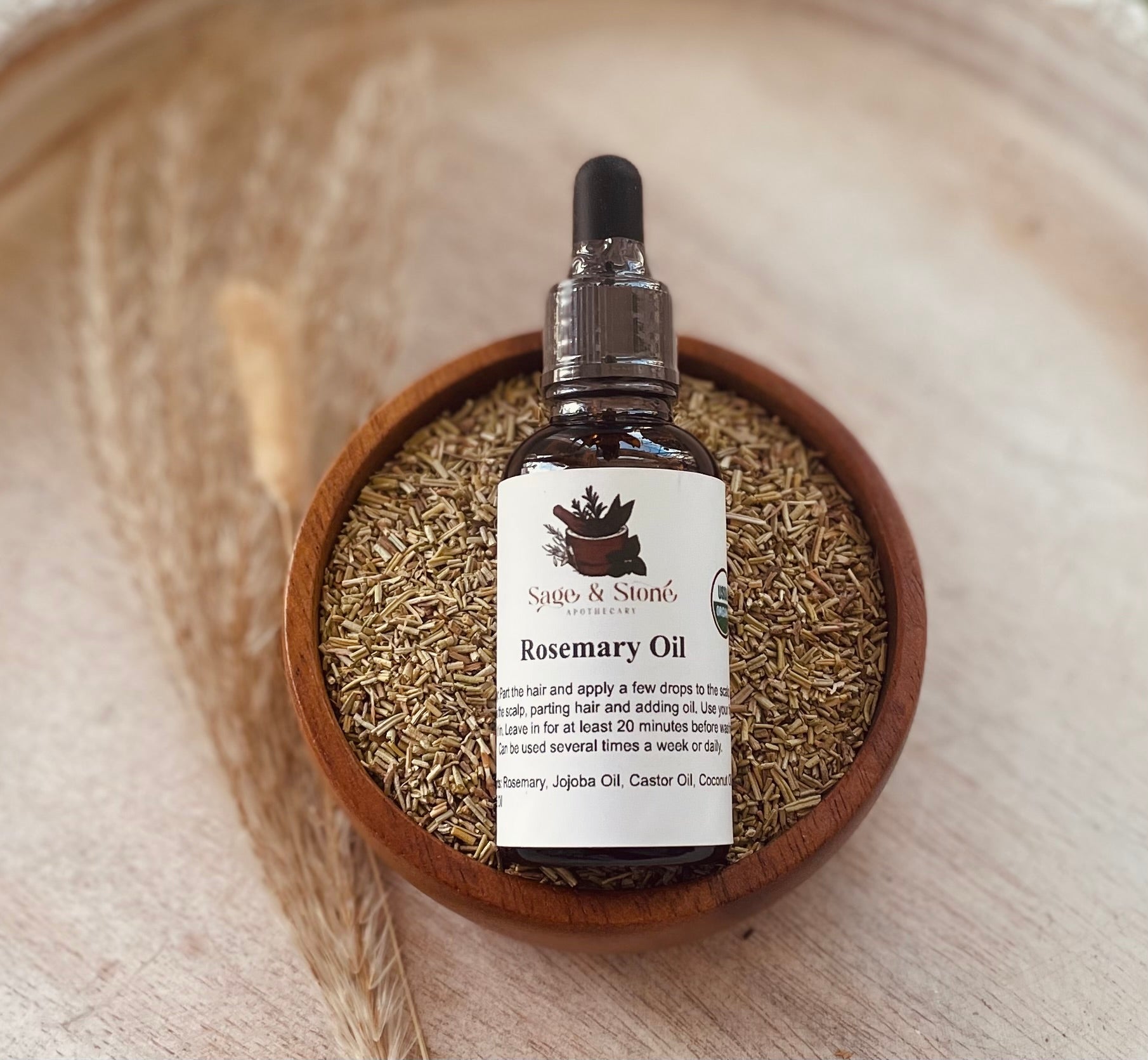 Bottle of Sage & Stone Apothecary Rosemary Oil for body care and wellness, with fresh rosemary sprigs beside it.