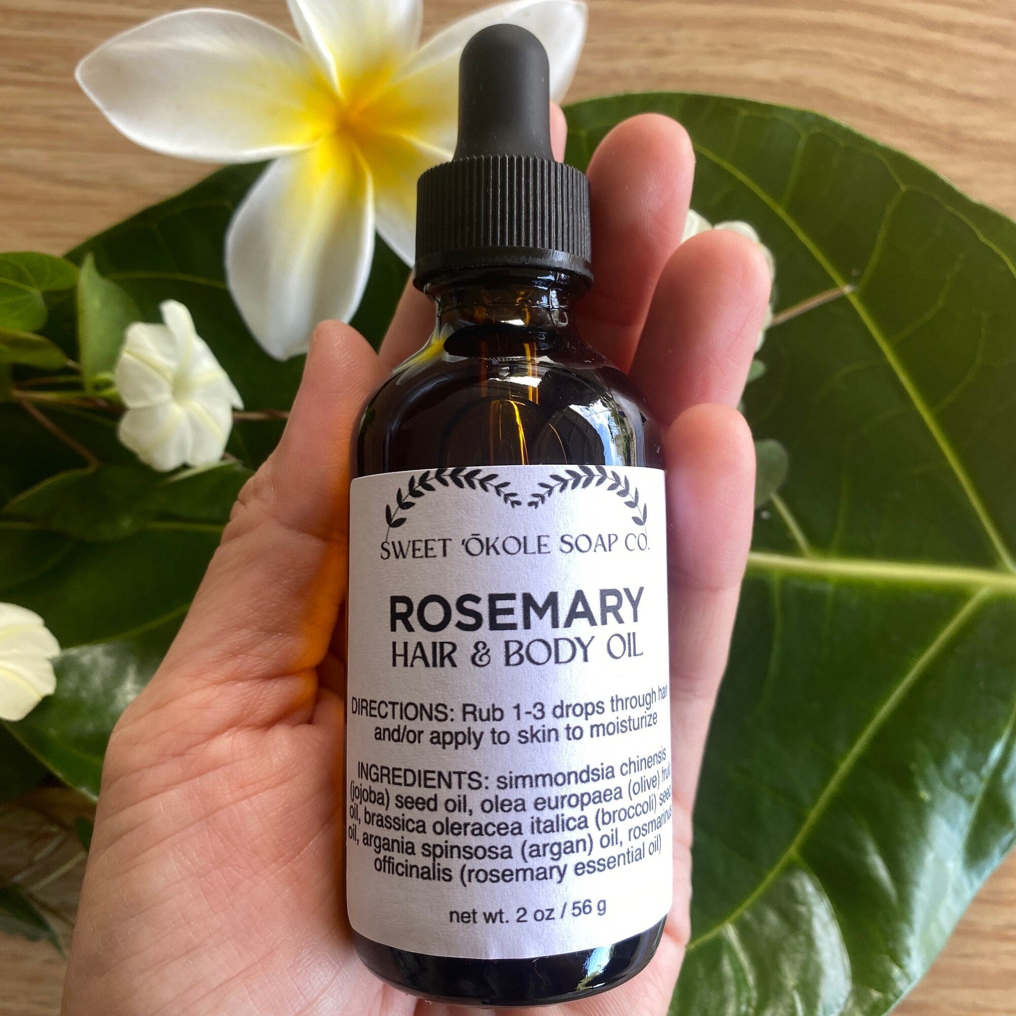 Bottle of Sweet ʻŌkole Soap Rosemary Hair & Scalp Oil with rosemary sprigs and oil in a clear glass container.