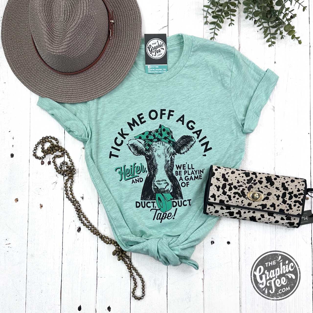 The Graphic Tee Tick Me Off Again Heifer Short Sleeve Heather Prism Mint graphic T-shirt showing a humorous design on the front.