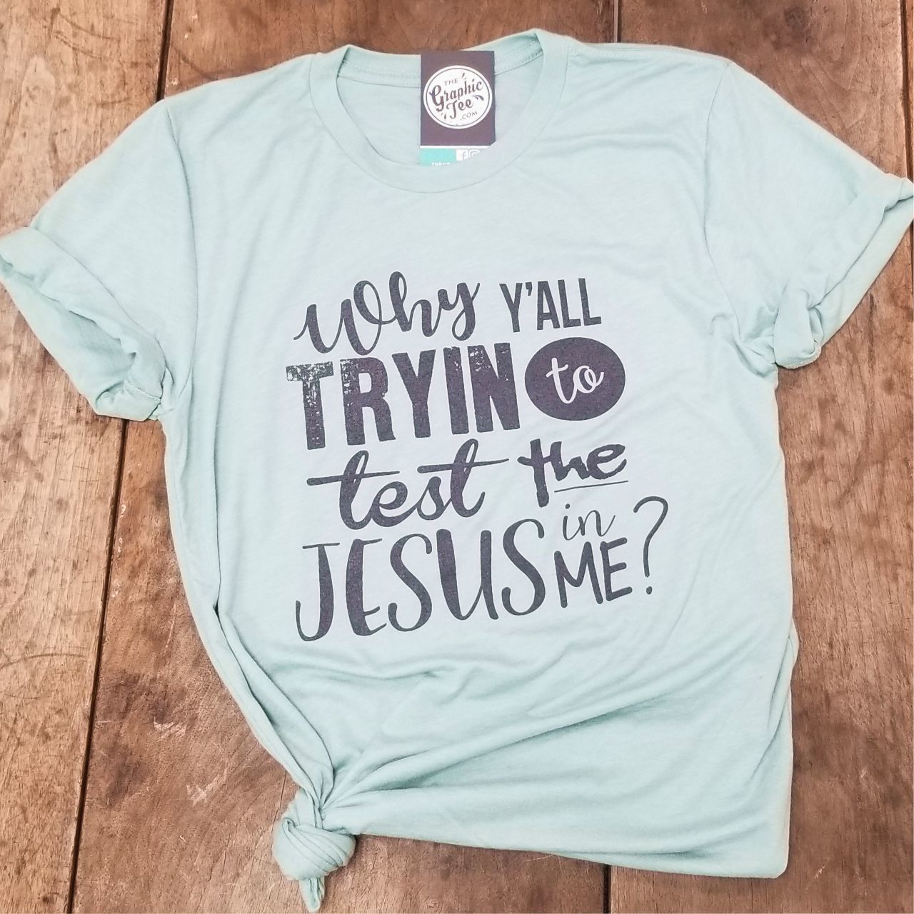 Dusty blue graphic t-shirt from The Graphic Tee featuring the phrase "Why Y'all Tryin to Test the Jesus in Me" in bold text.