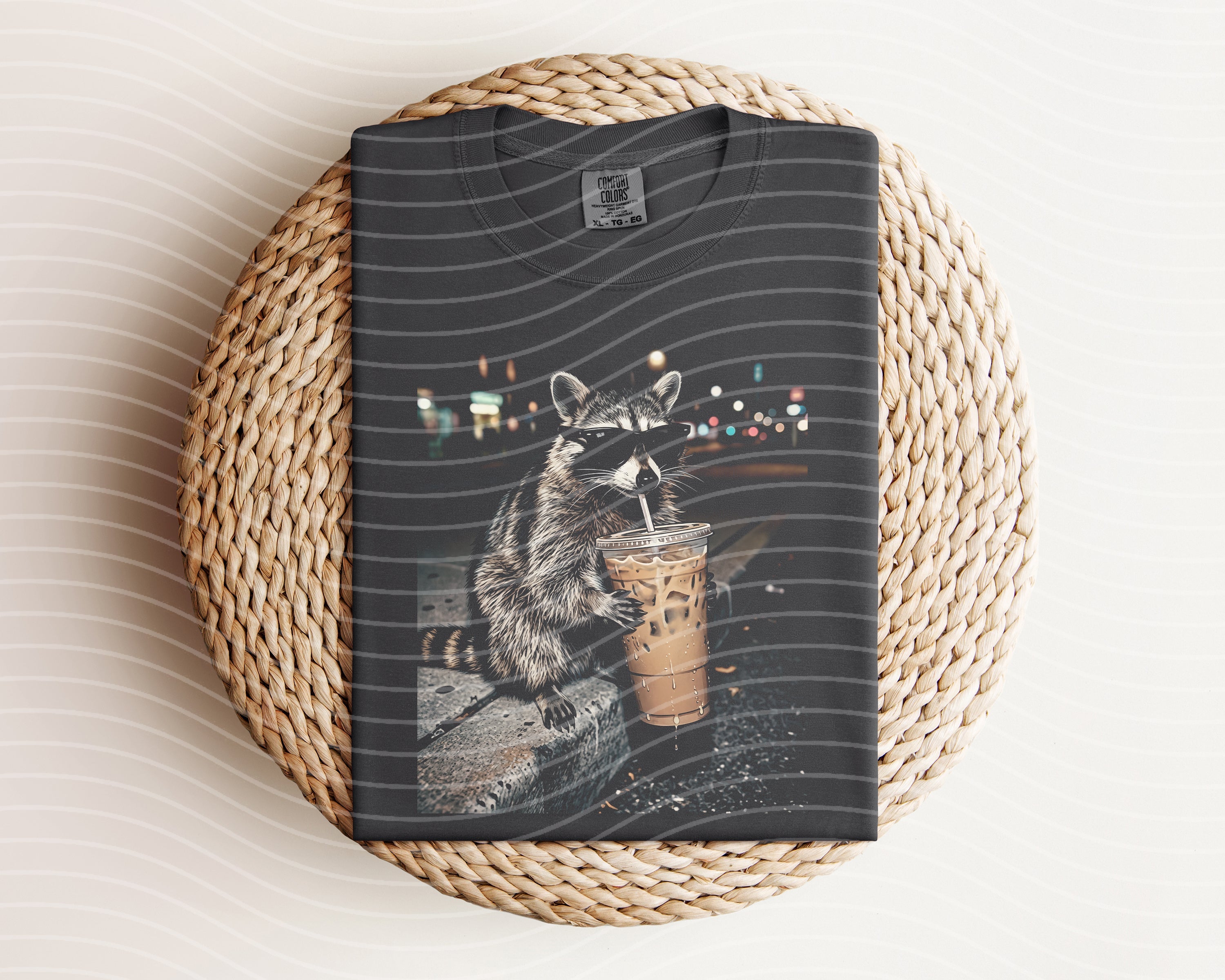 Iced coffee raccoon graphic tee from Anchored Grace Boutique, featuring a playful raccoon holding a coffee cup on soft fabric.