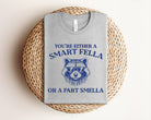 Graphic tee featuring humorous "You're Either a Smart Fella or a Fart Smella" design in black and white, from Anchored Grace Boutique.