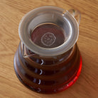 Clear glass two-cup teapot by August Uncommon Tea, showcasing a sleek design with a handle, spout, and lid for brewing tea.