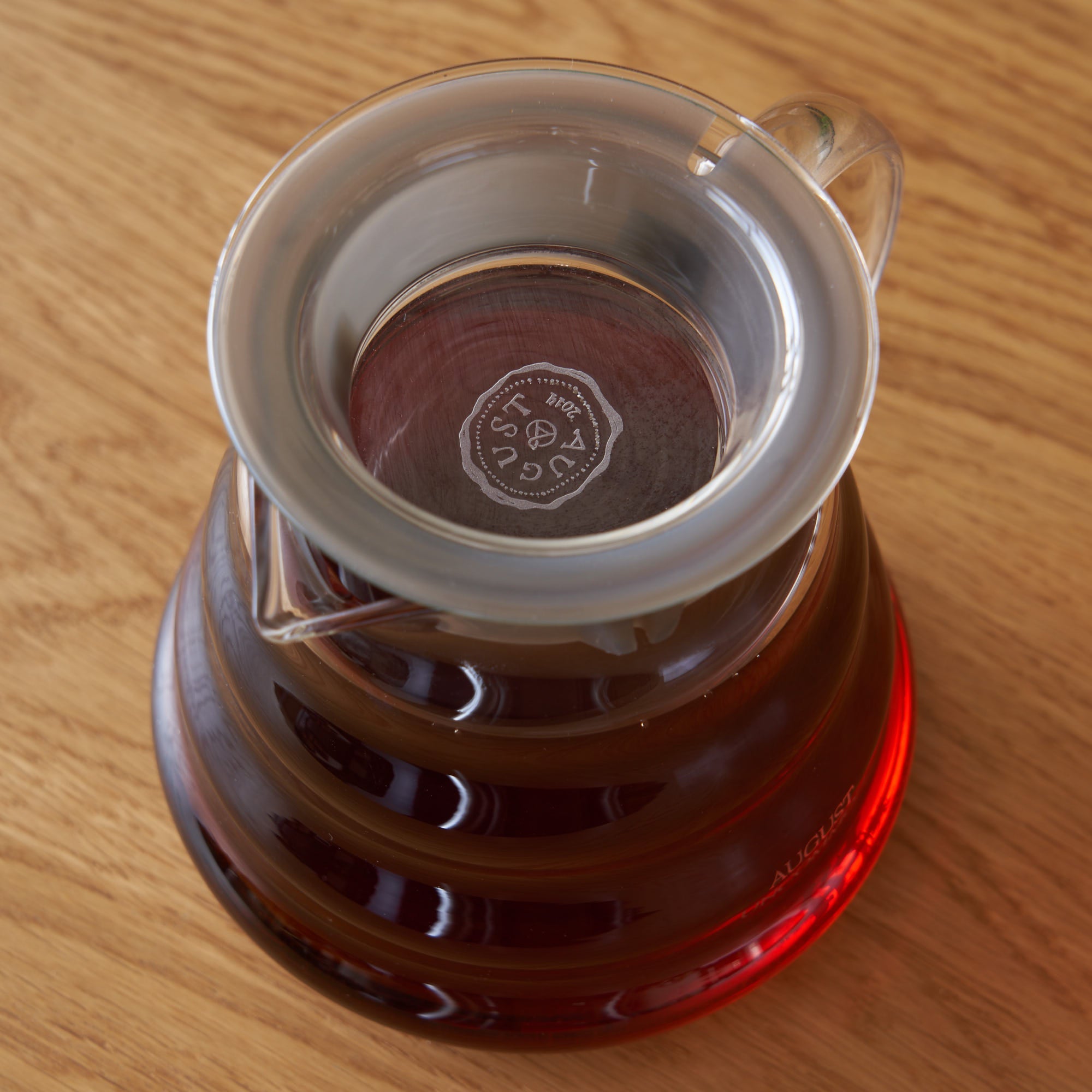 Clear glass two-cup teapot by August Uncommon Tea, showcasing a sleek design with a handle, spout, and lid for brewing tea.
