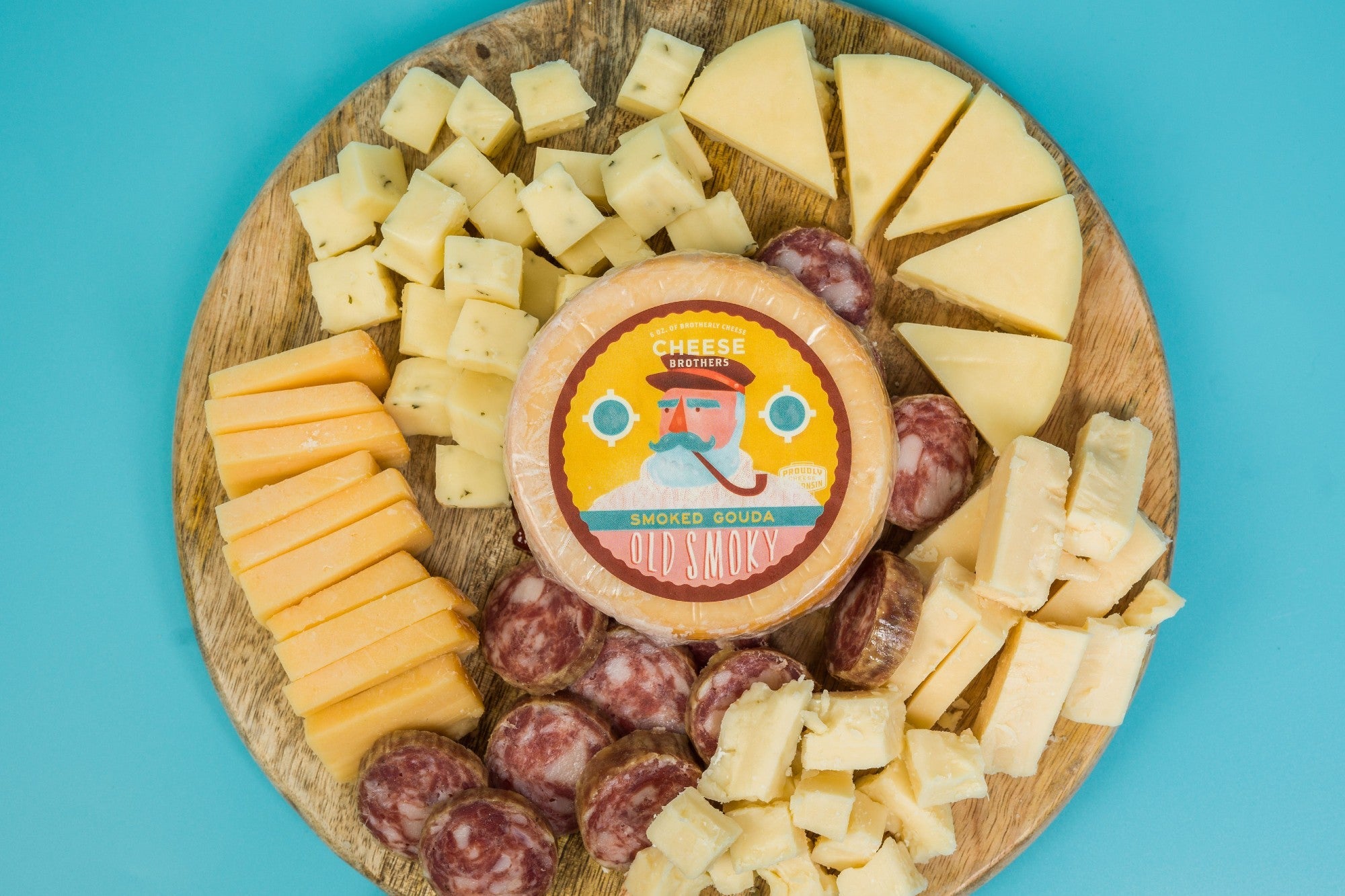 Assorted cheese and meat charcuterie pack from Cheese Brothers, Inc., arranged with crackers and fresh fruits on a wooden board.