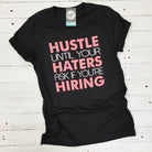 Black V-neck graphic T-shirt by The Graphic Tee with bold "Hustle Until Your Haters Ask If You're Hiring" design.