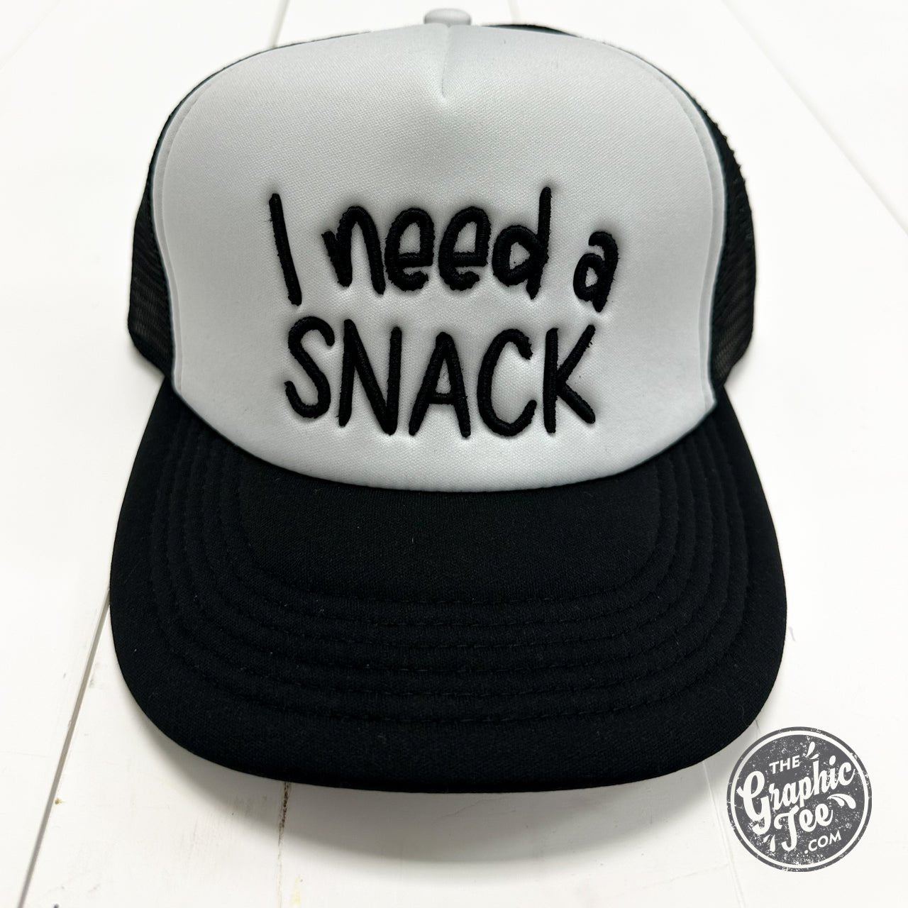 Foam trucker cap with "I Need A Snack" graphic by The Graphic Tee, featuring a structured brim and adjustable snapback closure.
