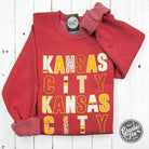 Kansas City multi-pattern letters on Heather Sport Scarlet crewneck sweatshirt by The Graphic Tee, featuring a casual, stylish design.