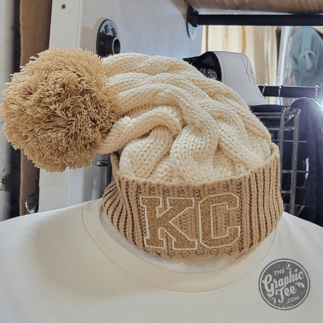 KC Glitter Applique Twist Knit Beanie with Pom, stylish cap featuring glitter accents and cozy knit material, by The Graphic Tee.