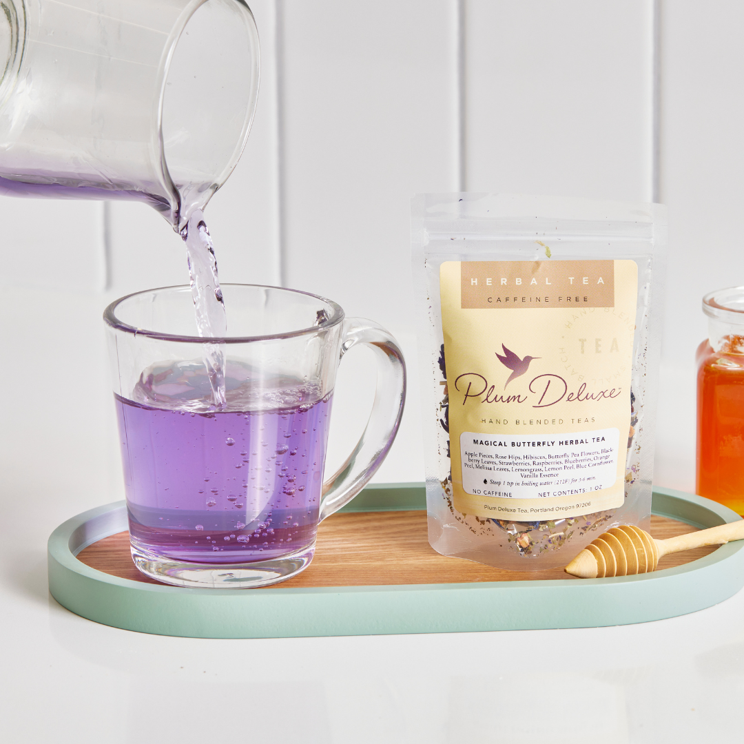Plum Deluxe Magical Butterfly Herbal Tea with butterfly pea flowers and berries, displayed in a clear glass teacup with flowers.