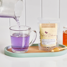 Plum Deluxe Magical Butterfly Herbal Tea with butterfly pea flowers and berries, displayed in a clear glass teacup with flowers.