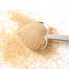 Plum Deluxe Honey Dust with cane sugar and raw honey, shown in a clear jar with a scoop on a rustic wooden surface.