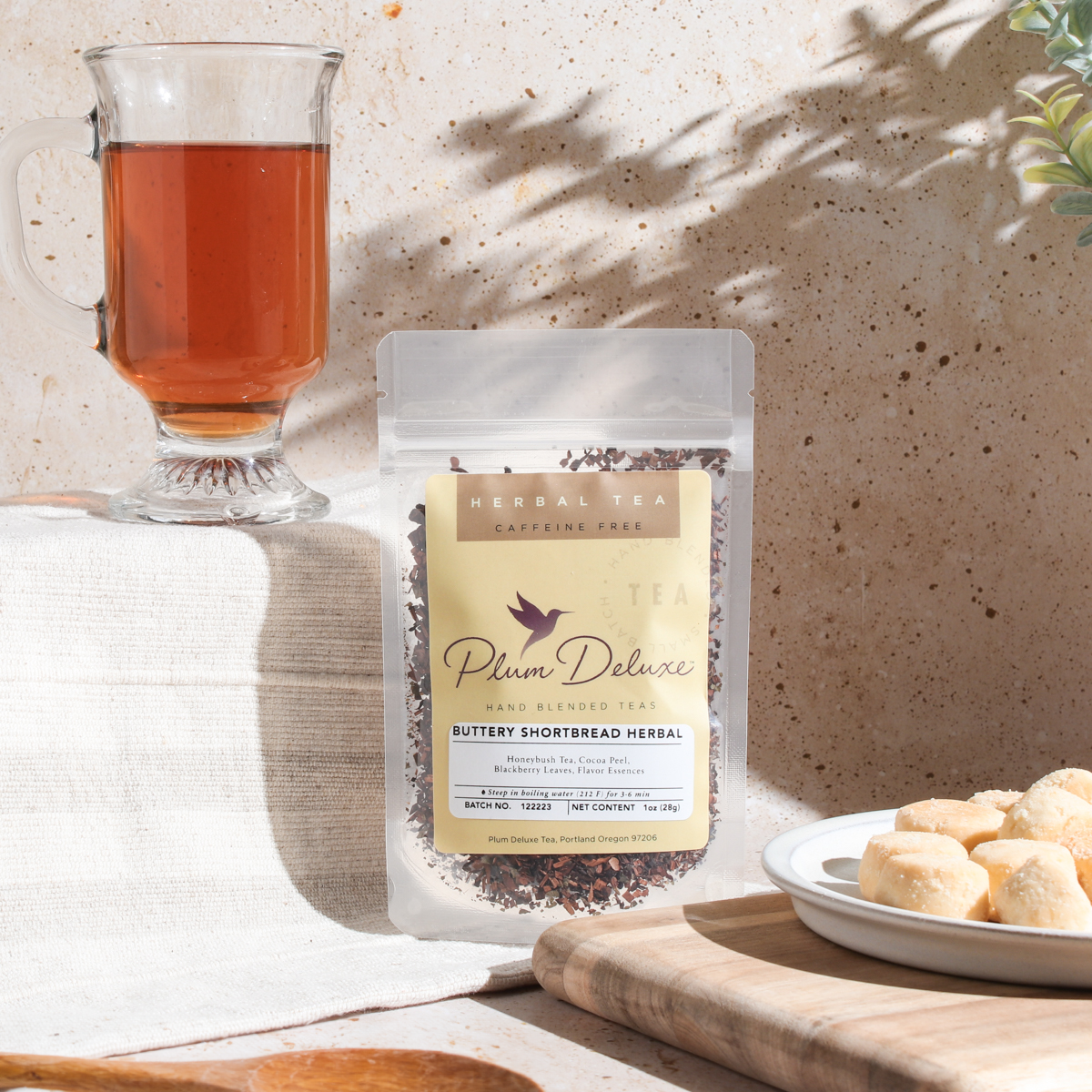 Plum Deluxe Tea Buttery Shortbread Herbal Tea in a clear glass cup with a tea infuser and fresh herbs on a white background.