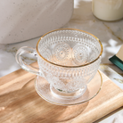 Clear glass teacup with matching saucer from Plum Deluxe Tea's Café Society collection, perfect for enjoying hot tea.