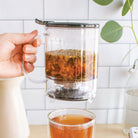 Plum Deluxe Tea Perfect Tea Maker brewing loose leaf tea in a glass infuser with a handle and lid, on a kitchen countertop.