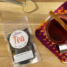 A cup of Vanilla Toddy Black Tea by Plum Deluxe Tea with vanilla beans and black tea leaves on a cozy table setting.