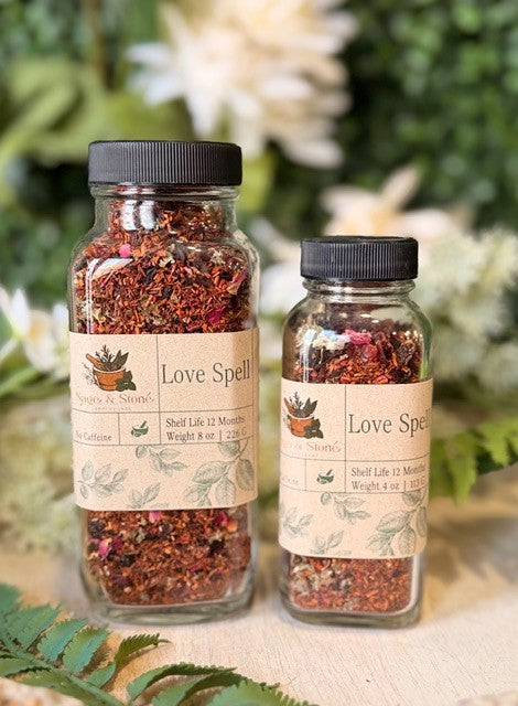 Sage & Stone Apothecary Love Spell Herbal Tea in a clear glass teapot with blooming herbs and a cozy setting.