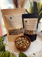 A cup of Moon Goddess Herbal Tea from Sage & Stone Apothecary with dried herbs and tea leaves on a rustic wooden surface.