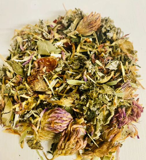 A cup of Sage & Stone Apothecary Nourishing Pregnancy Tea in a glass mug with fresh herbs and tea leaves on a wooden table.