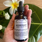 Bottle of Sweet ʻŌkole Soap Rosemary Hair & Scalp Oil with rosemary sprigs and oil in a clear glass container.