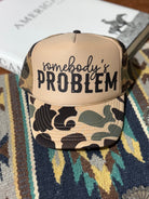 Wild Junkie Somebody's Problem camo trucker hat with mesh back and curved brim, featuring a bold embroidered design.